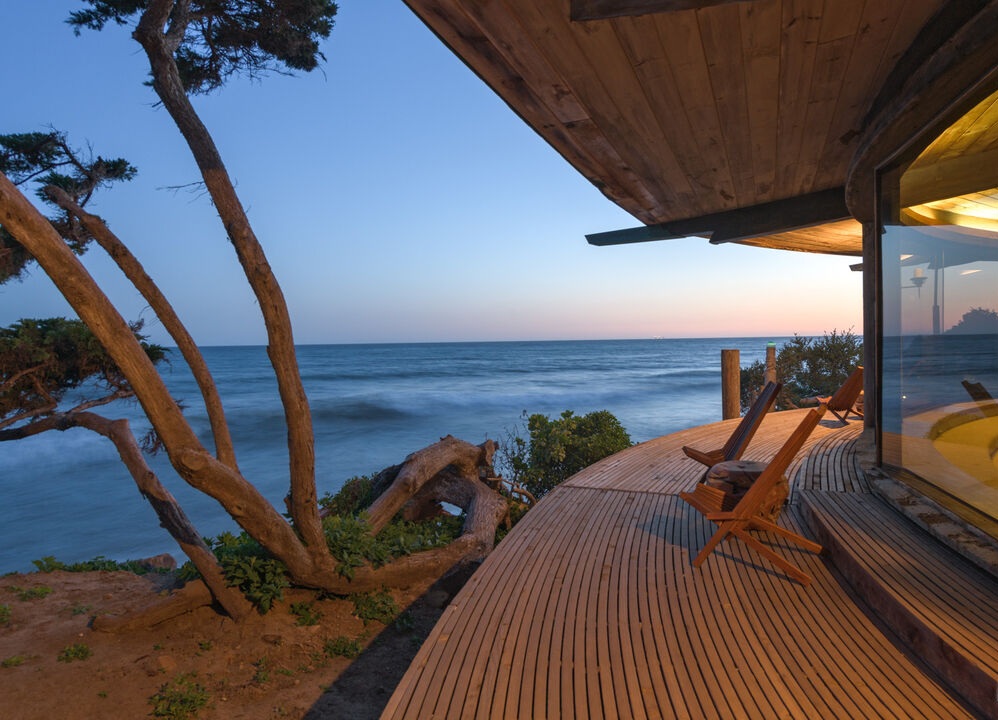 Harry Gesner’s Malibu Sandcastle Home at late sunset with view of the wrap around deck and Pacific Ocean in the background and unique cypress flora elements and beach stones and drift wood in the forefront.